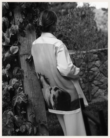 Woman in white shirt with horse print poses against a wooden post outdoors amid trees and plants in a black-and-white fashion shot.