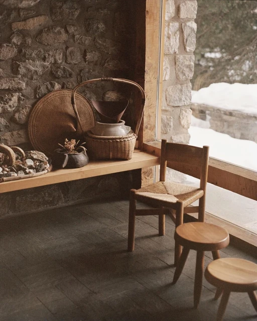 Rustic interior with stone walls, wooden furniture, baskets, and view to snow-covered ground outside the window, capturing a cozy, natural aesthetic.