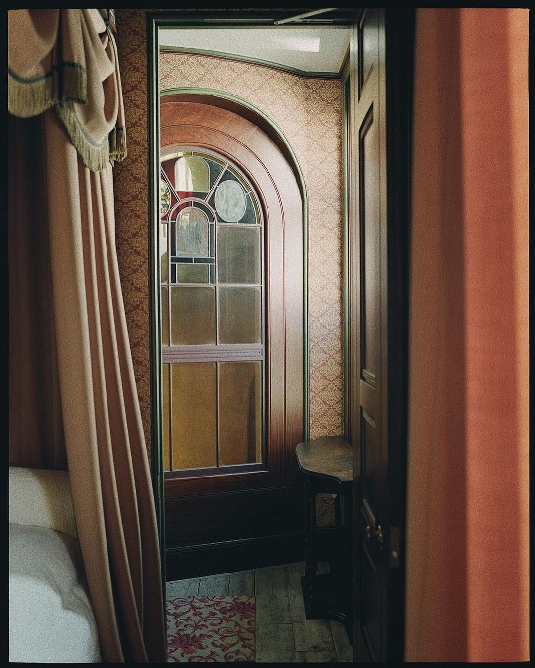 Interior view showcases arched stained-glass window, vintage wallpaper, and cozy bedroom ambiance with a curtained four-poster bed.