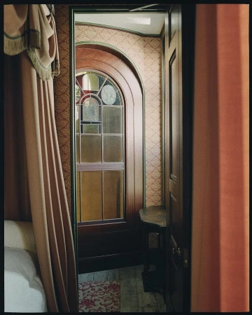 Interior view showcases arched stained-glass window, vintage wallpaper, and cozy bedroom ambiance with a curtained four-poster bed.