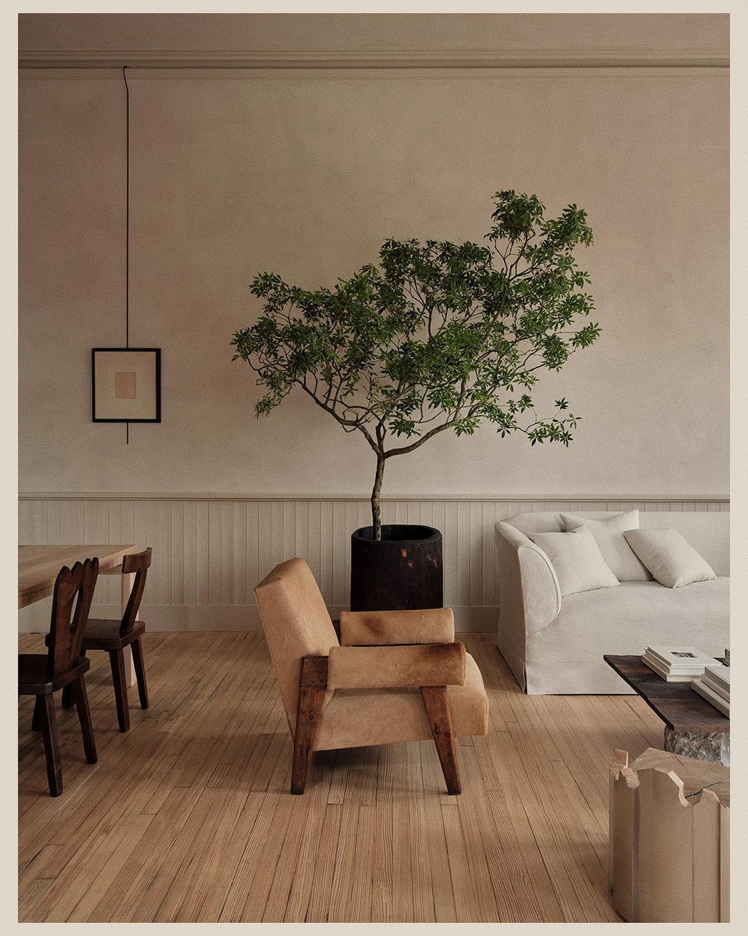 Living room featuring a neutral-toned armchair, potted tree, white couch, wooden floor, and natural light, creating a warm and inviting atmosphere.