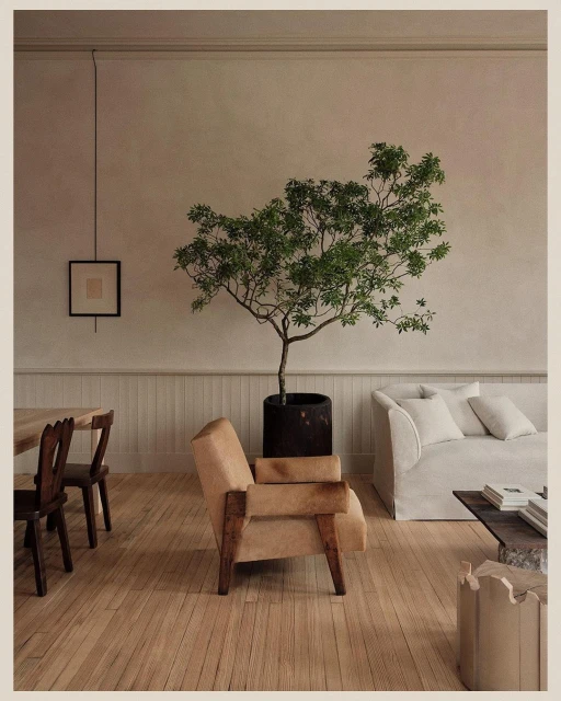 Living room featuring a neutral-toned armchair, potted tree, white couch, wooden floor, and natural light, creating a warm and inviting atmosphere.