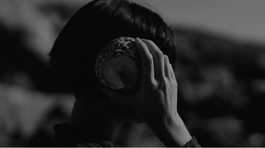 Monochrome shot of an adult holding a textured, spherical object to their ear with a blurred outdoor background.