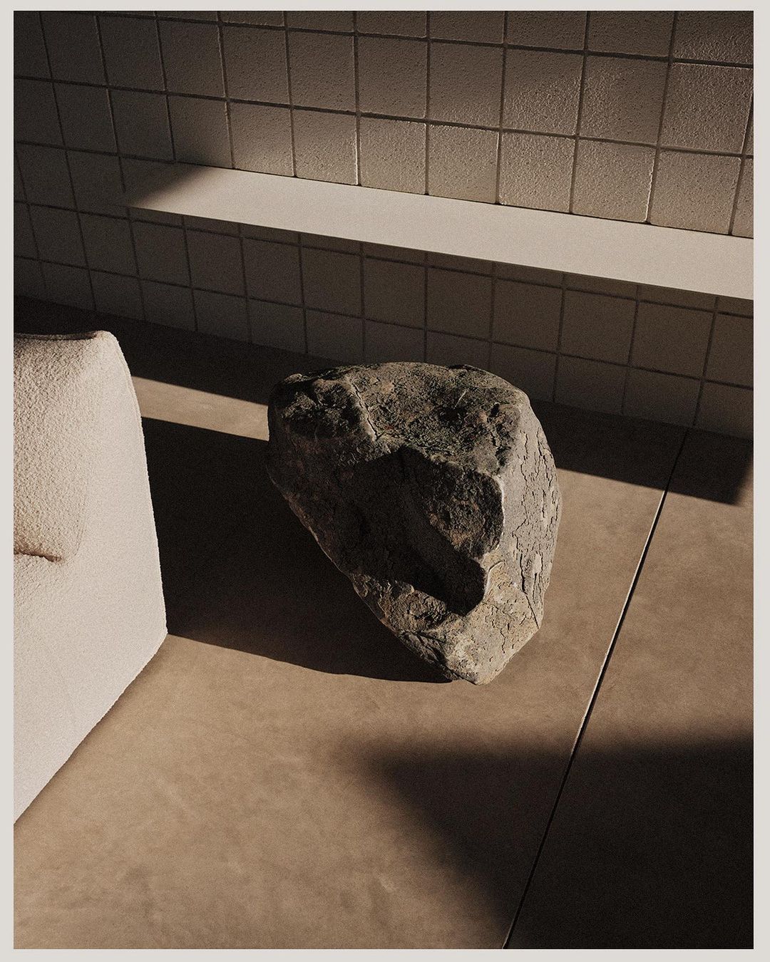 Minimalist interior still life with a large grey stone sitting on a tan floor near a textured off-white couch and tiled wall with sunlight.