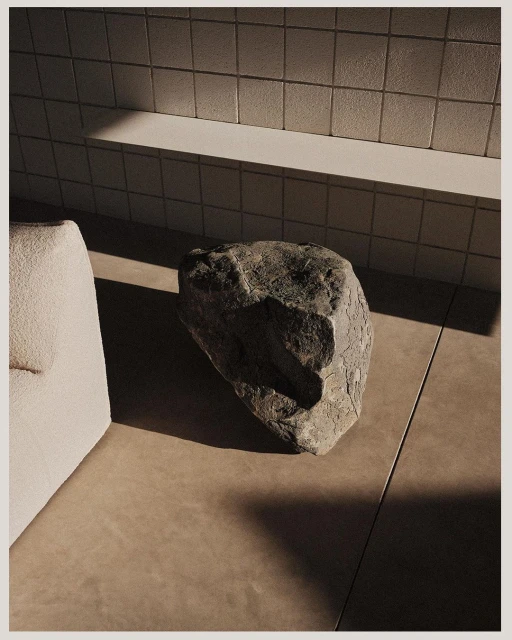 Minimalist interior still life with a large grey stone sitting on a tan floor near a textured off-white couch and tiled wall with sunlight.