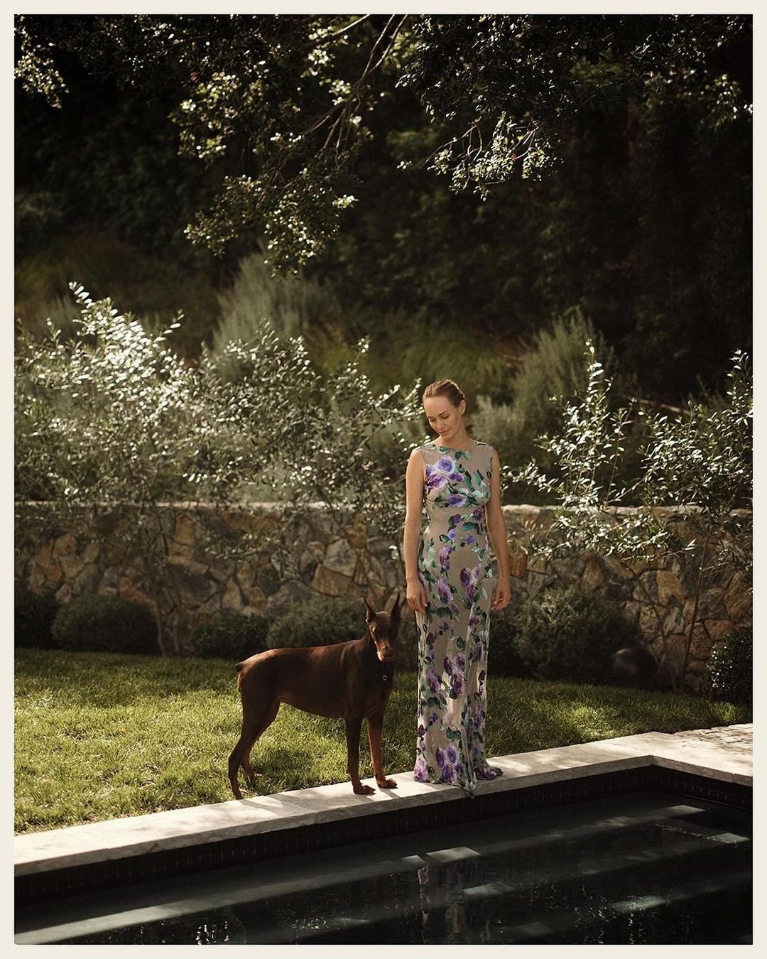 An adult woman stands with a brown Doberman dog alongside a swimming pool in a backyard with a stone wall and overgrown bushes.