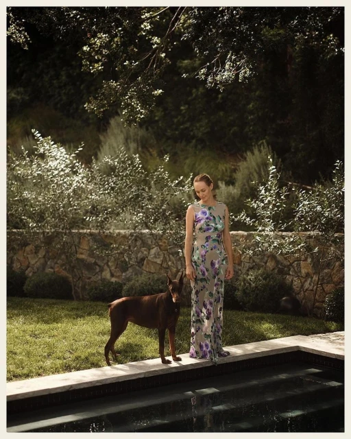 An adult woman stands with a brown Doberman dog alongside a swimming pool in a backyard with a stone wall and overgrown bushes.