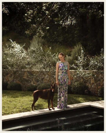 An adult woman stands with a brown Doberman dog alongside a swimming pool in a backyard with a stone wall and overgrown bushes.