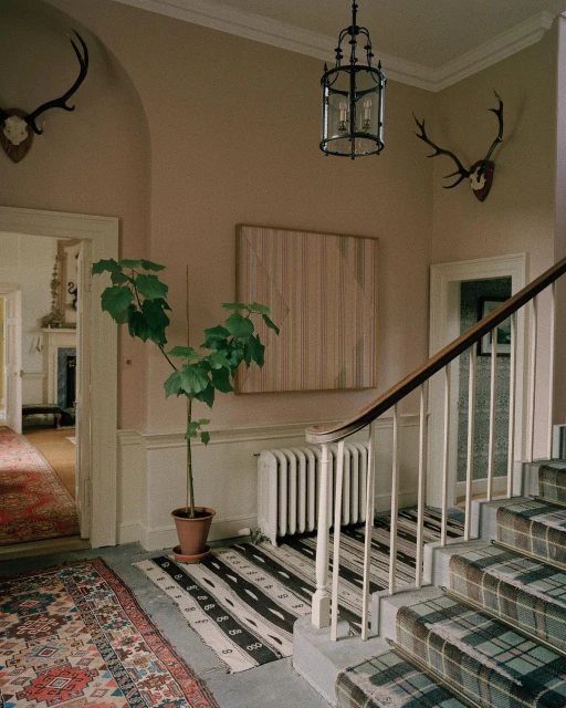 Interior of a hallway with staircase covered in plaid, potted plant, striped rug, antique antlers on the wall, and decorative light fixture.