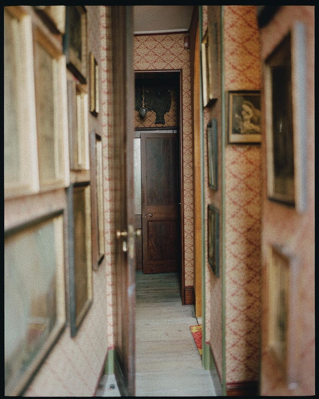Interior hallway with framed art on patterned walls leading to a dark wood door and decorated wall. The shot is slightly blurred.