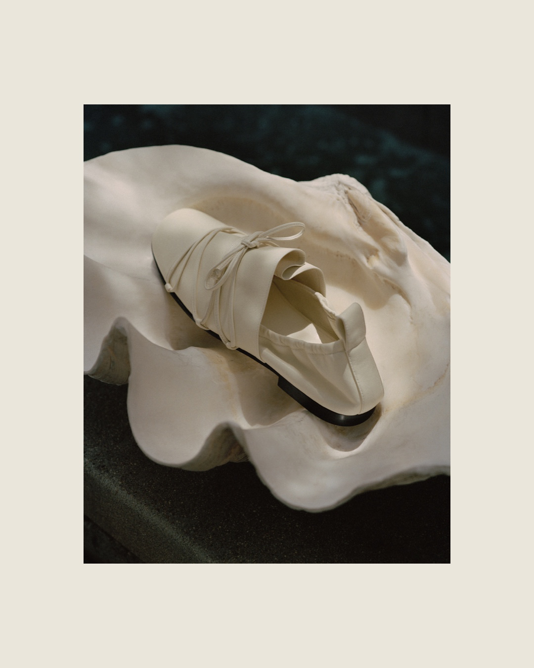 Still life of white ballet flat shoe, featuring ruffled upper, displayed on a white clam shell against a dark green background.