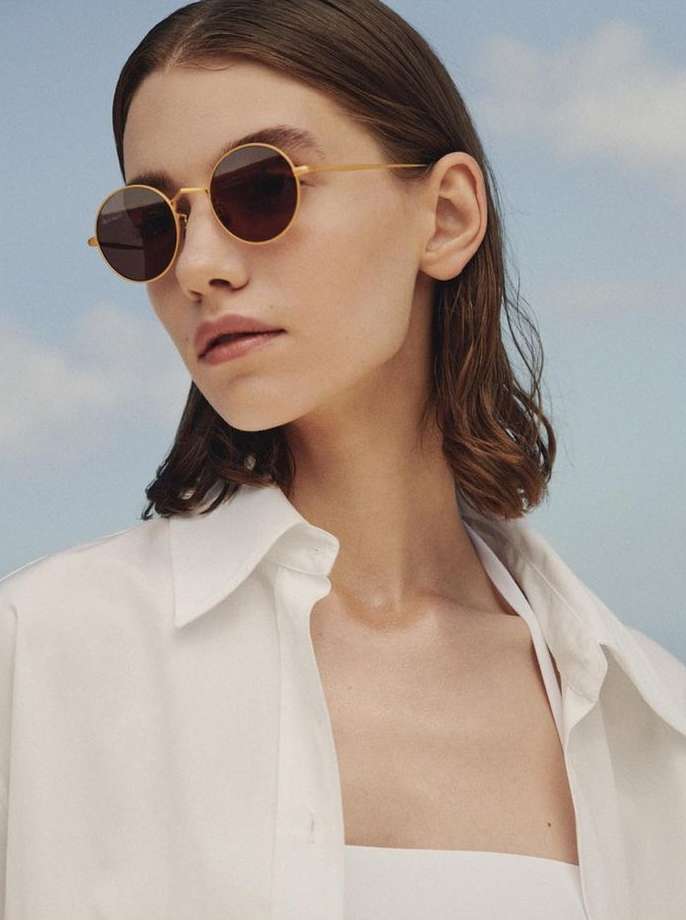 Close-up of a female model with round, gold-rimmed sunglasses, fair skin, and wavy dark hair, wearing a white collared shirt against a blue sky.