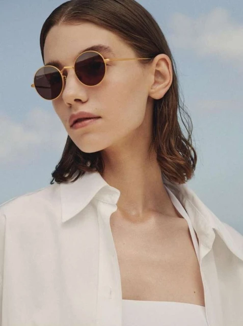 Close-up of a female model with round, gold-rimmed sunglasses, fair skin, and wavy dark hair, wearing a white collared shirt against a blue sky.