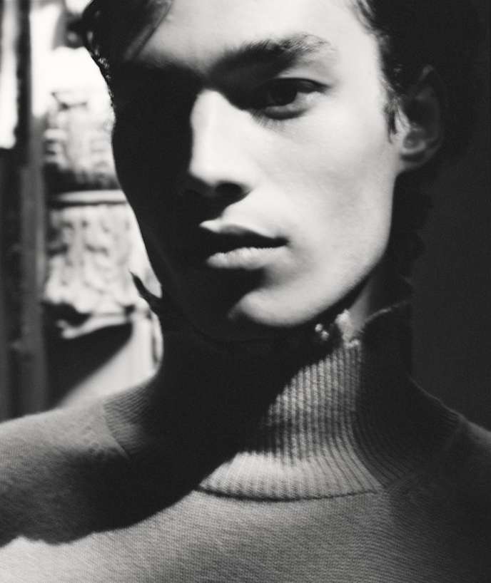 Close-up monochrome portrait of a young man wearing a high-neck sweater with dramatic high-contrast lighting.