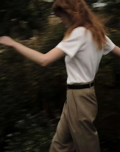 Woman with red hair wearing a white shirt and khaki pants, walking outside. The image has a shallow depth of field.