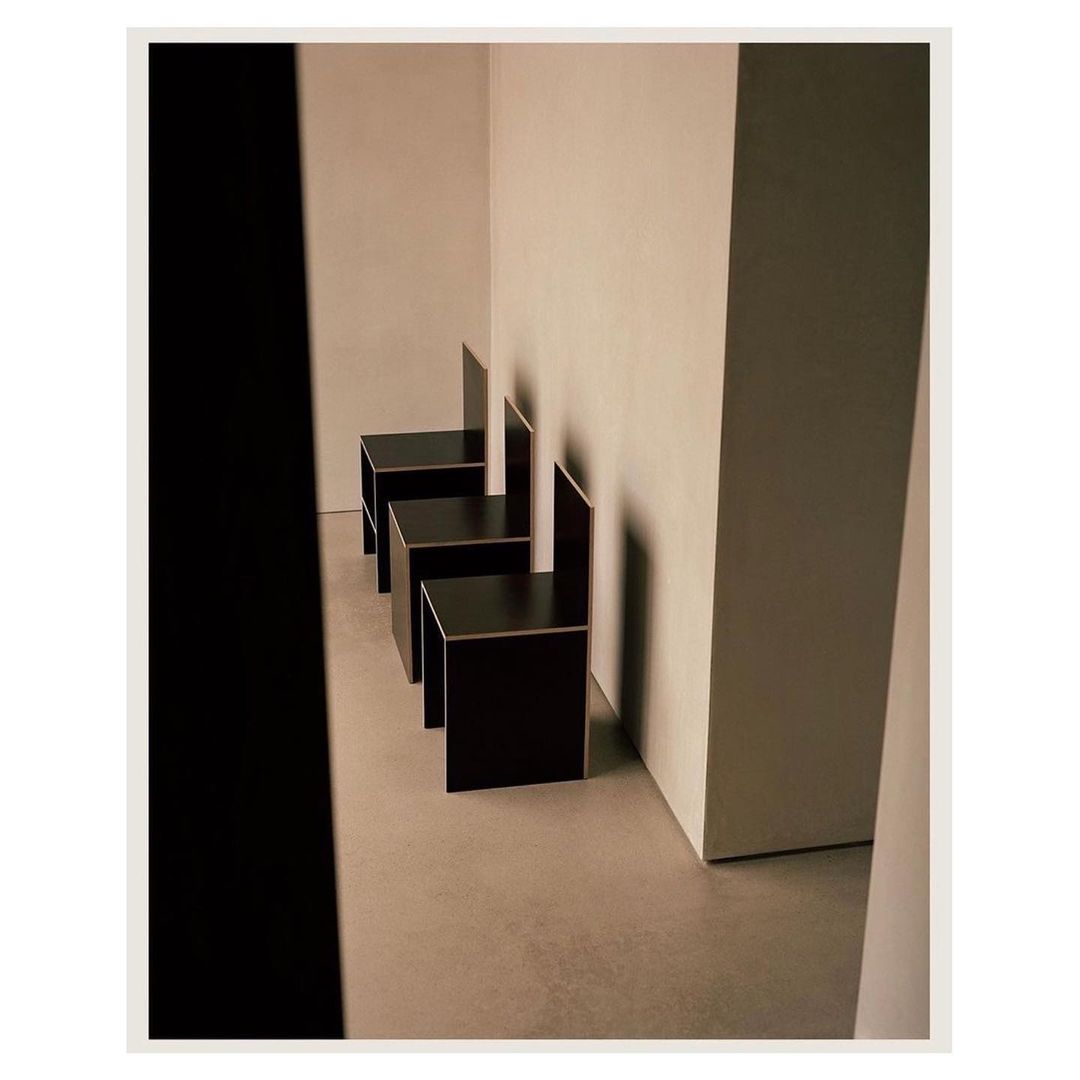 Three dark brown chairs with high backs arranged in a row in an off-white room, casting shadows on the walls and floor.