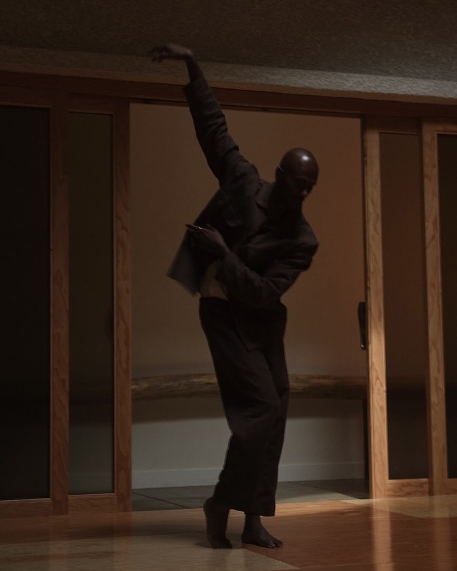 Adult male in suit dances barefoot inside of a home with wood floors and wood trimmed sliding glass doors in a medium indoor shot.