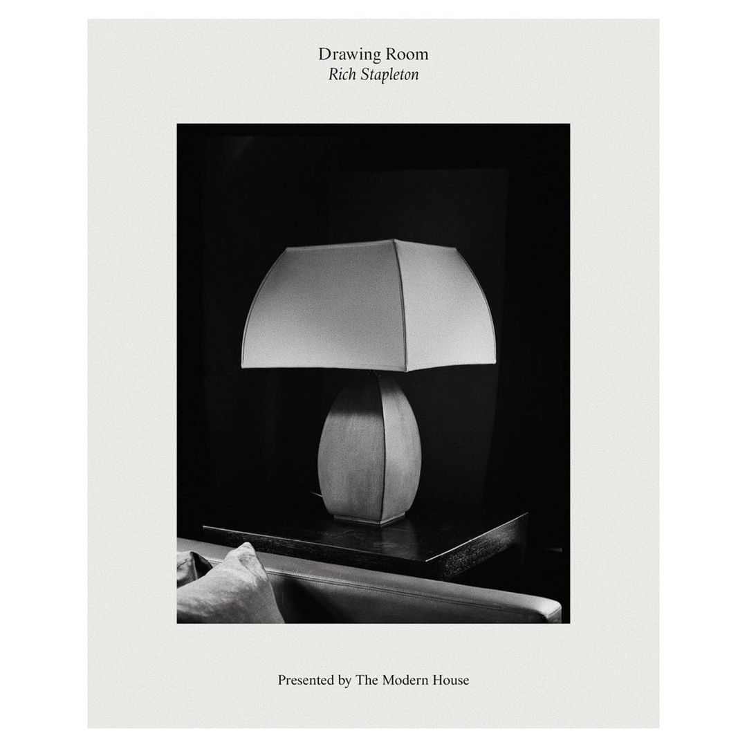 Black and white photograph of a lit table lamp with a square shade sitting on a dark surface, with a dark background.