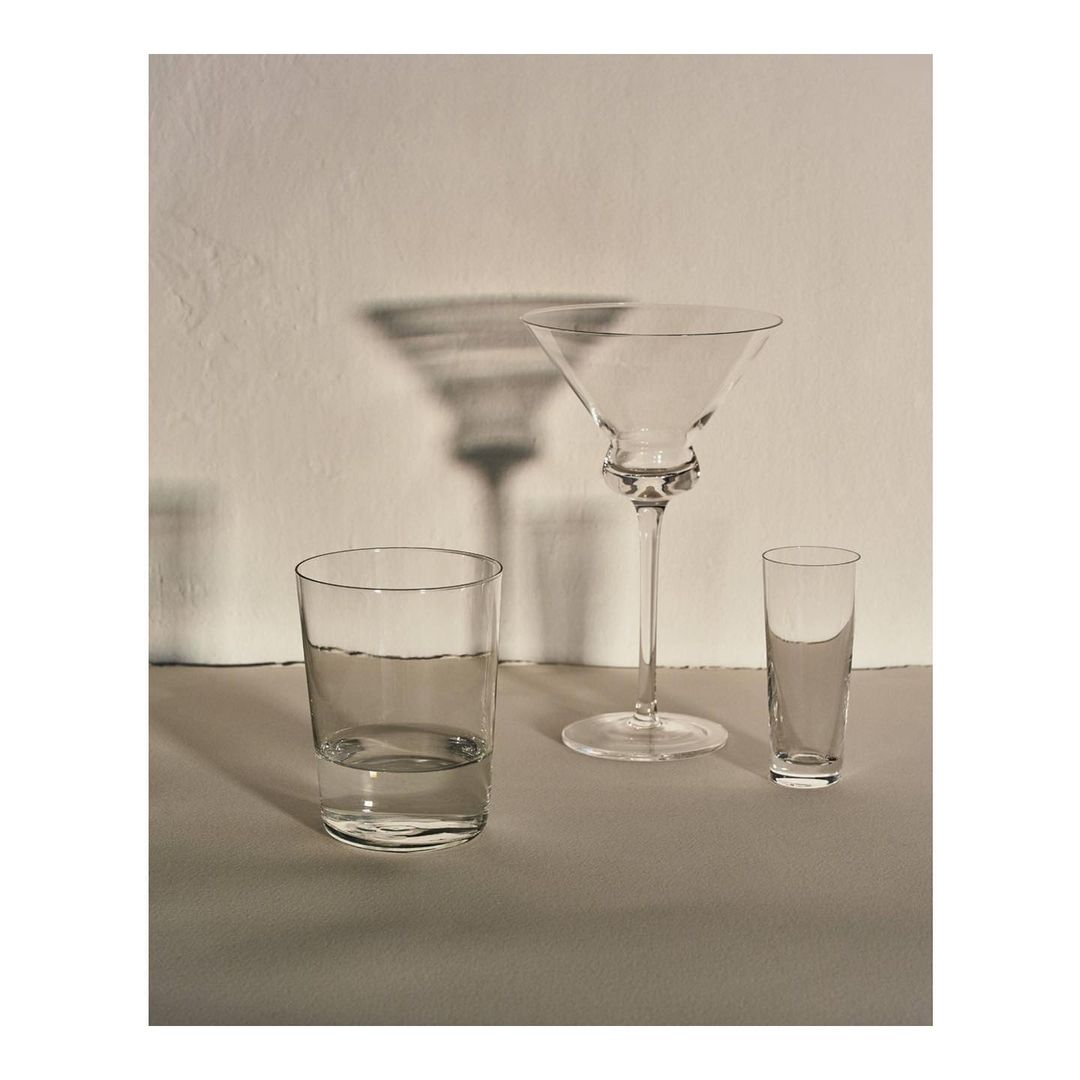 Elegant still life features a martini glass, drinking glass, and shot glass on a reflective surface against a neutral background, casting soft shadows.