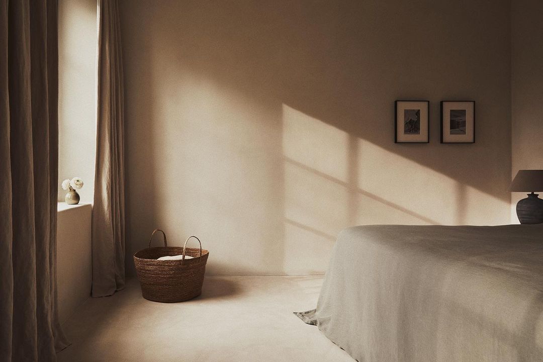 Bedroom interior with natural light, linen bedding on bed, woven basket, neutral color palette, and framed artwork on wall.