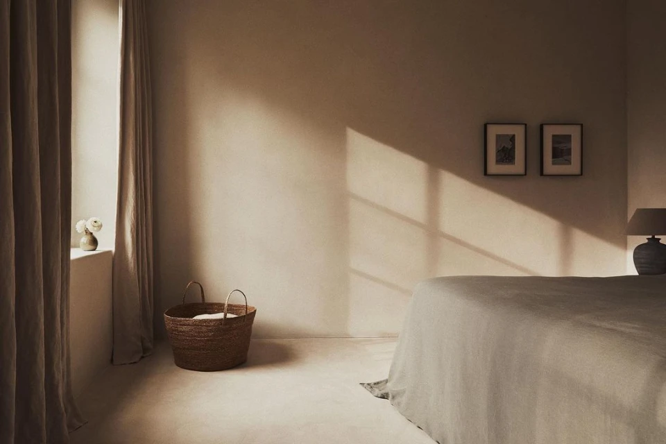 Bedroom interior with natural light, linen bedding on bed, woven basket, neutral color palette, and framed artwork on wall.