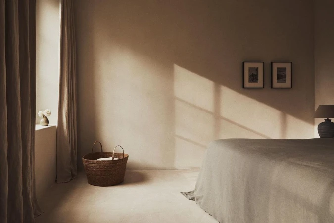 Bedroom interior with natural light, linen bedding on bed, woven basket, neutral color palette, and framed artwork on wall.