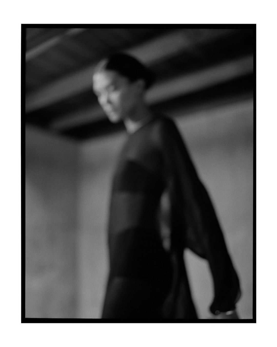 Monochrome portrait of an adult female in a long-sleeved dress, posing indoors with soft lighting and a shallow depth of field creating a blurred effect.
