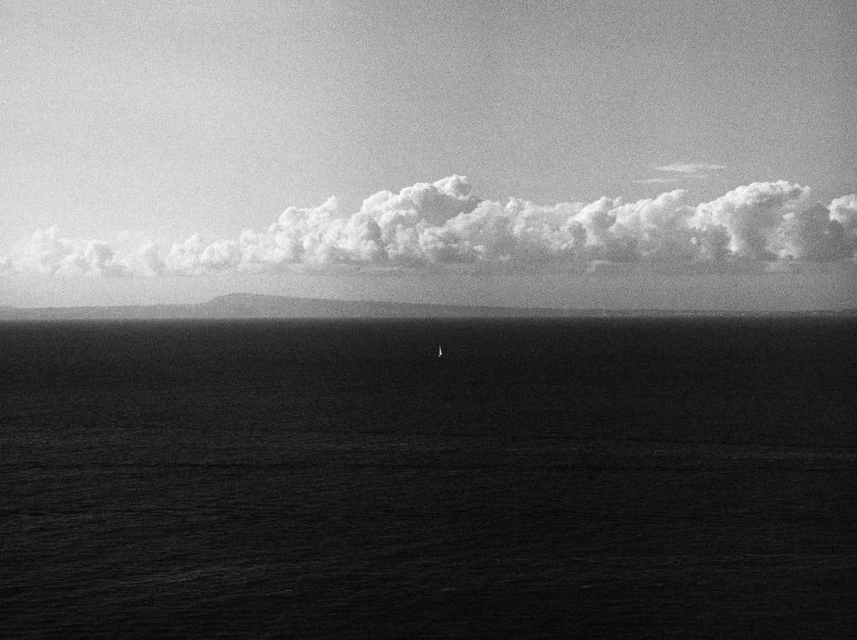 Monochrome seascape featuring a sailboat on the water, a distant landmass, and a sky filled with cumulus clouds.