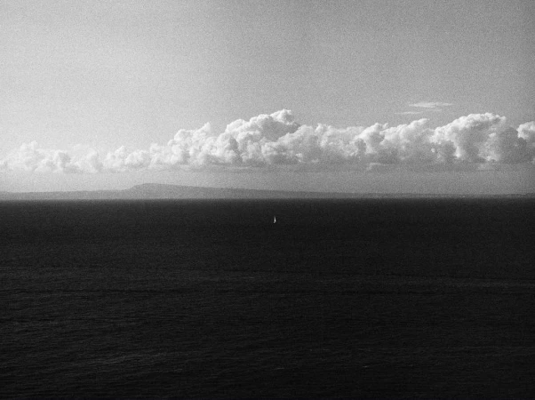Monochrome seascape featuring a sailboat on the water, a distant landmass, and a sky filled with cumulus clouds.