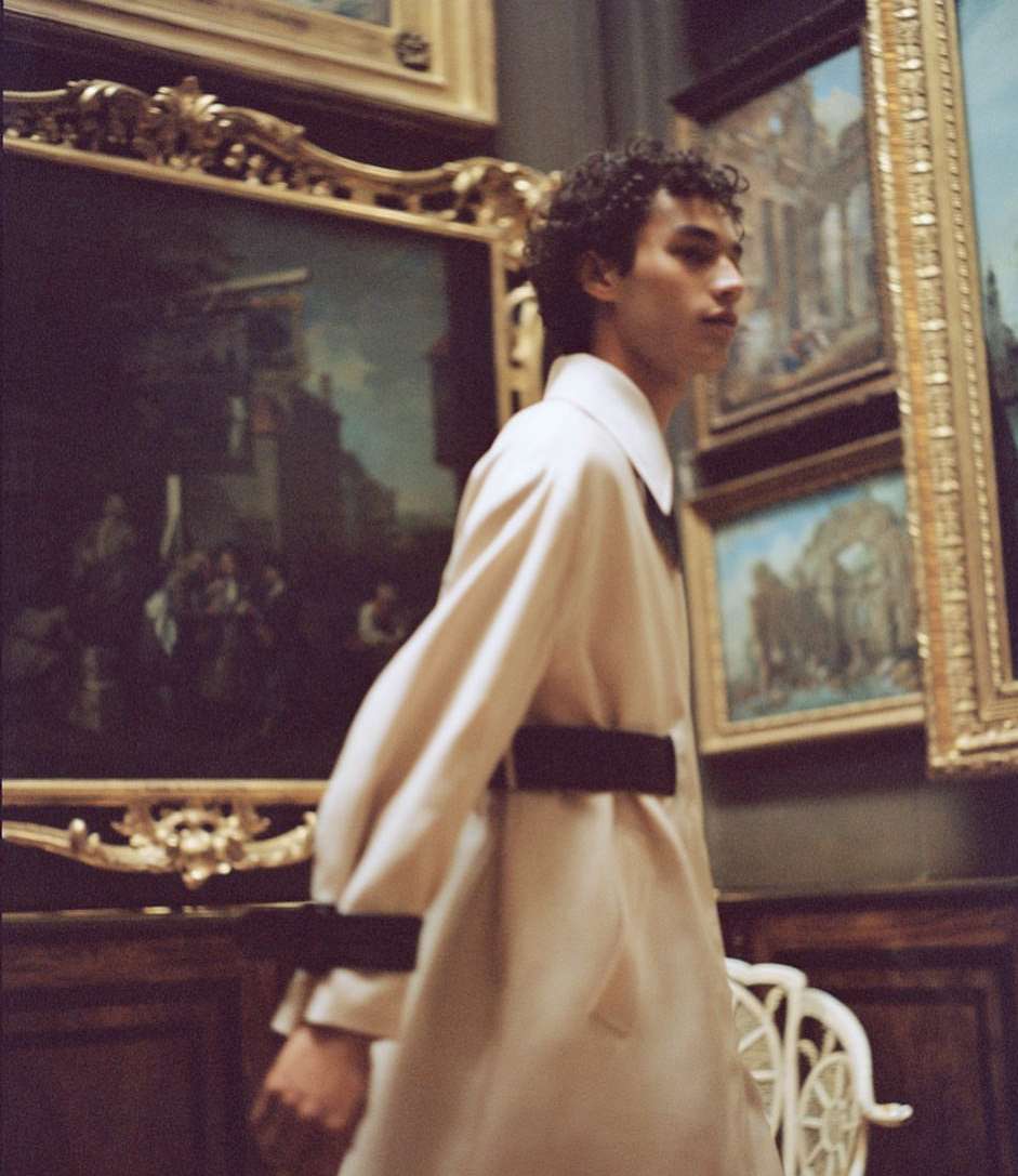 Young man in a beige coat, standing in an art gallery with multiple framed paintings on the wall and an ornate white chair nearby.