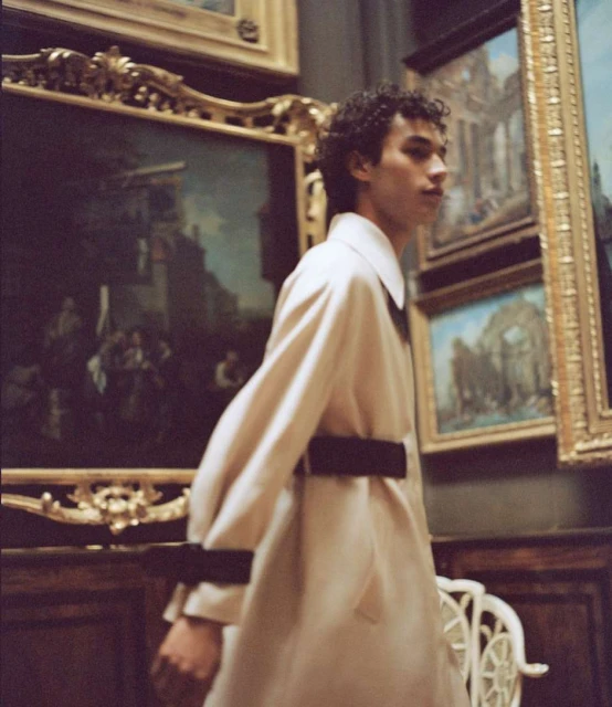 Young man in a beige coat, standing in an art gallery with multiple framed paintings on the wall and an ornate white chair nearby.