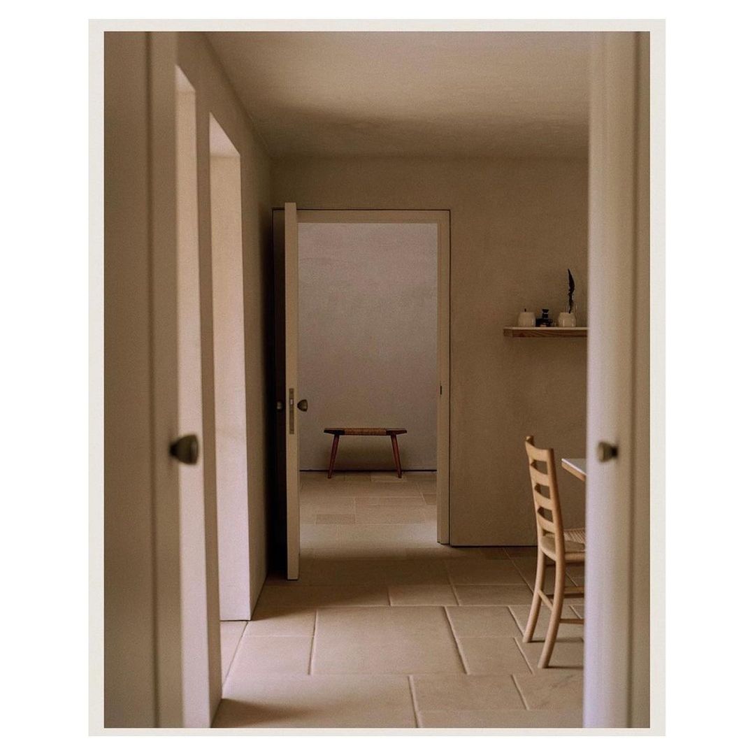 Indoor architectural shot through a doorway, revealing a bench in a sunlit room with beige walls and tile floors, plus a chair.