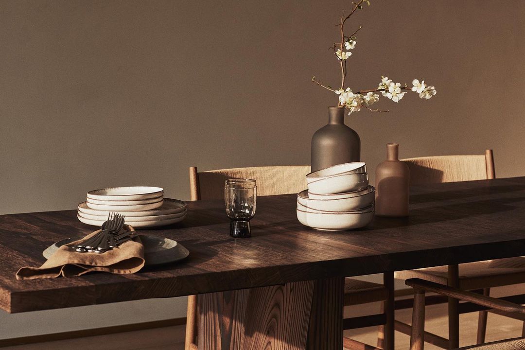 Dining table set with plates, bowls, glassware, flatware, linen napkin, and a vase with white blossoms on a wooden surface.