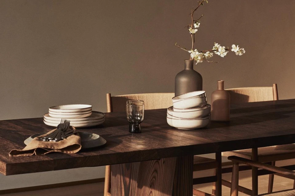 Dining table set with plates, bowls, glassware, flatware, linen napkin, and a vase with white blossoms on a wooden surface.