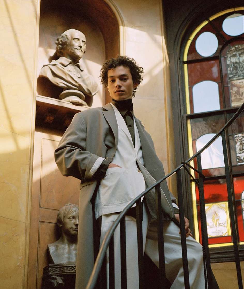 Fashionable young adult man poses on stairs next to bust sculptures, wearing a gray trench coat, vest, black turtleneck, and striped trousers.