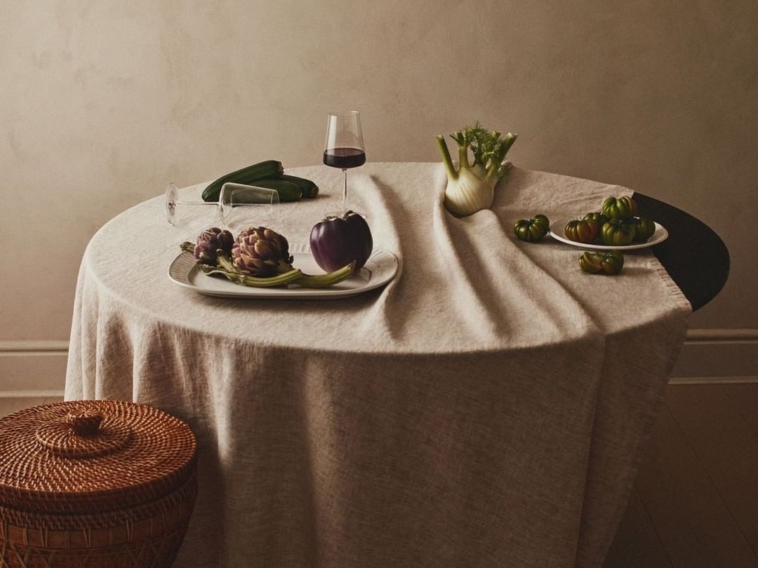 Still life on a round table with various vegetables, including artichokes, tomatoes, fennel, and cucumbers, plus a glass of red wine.