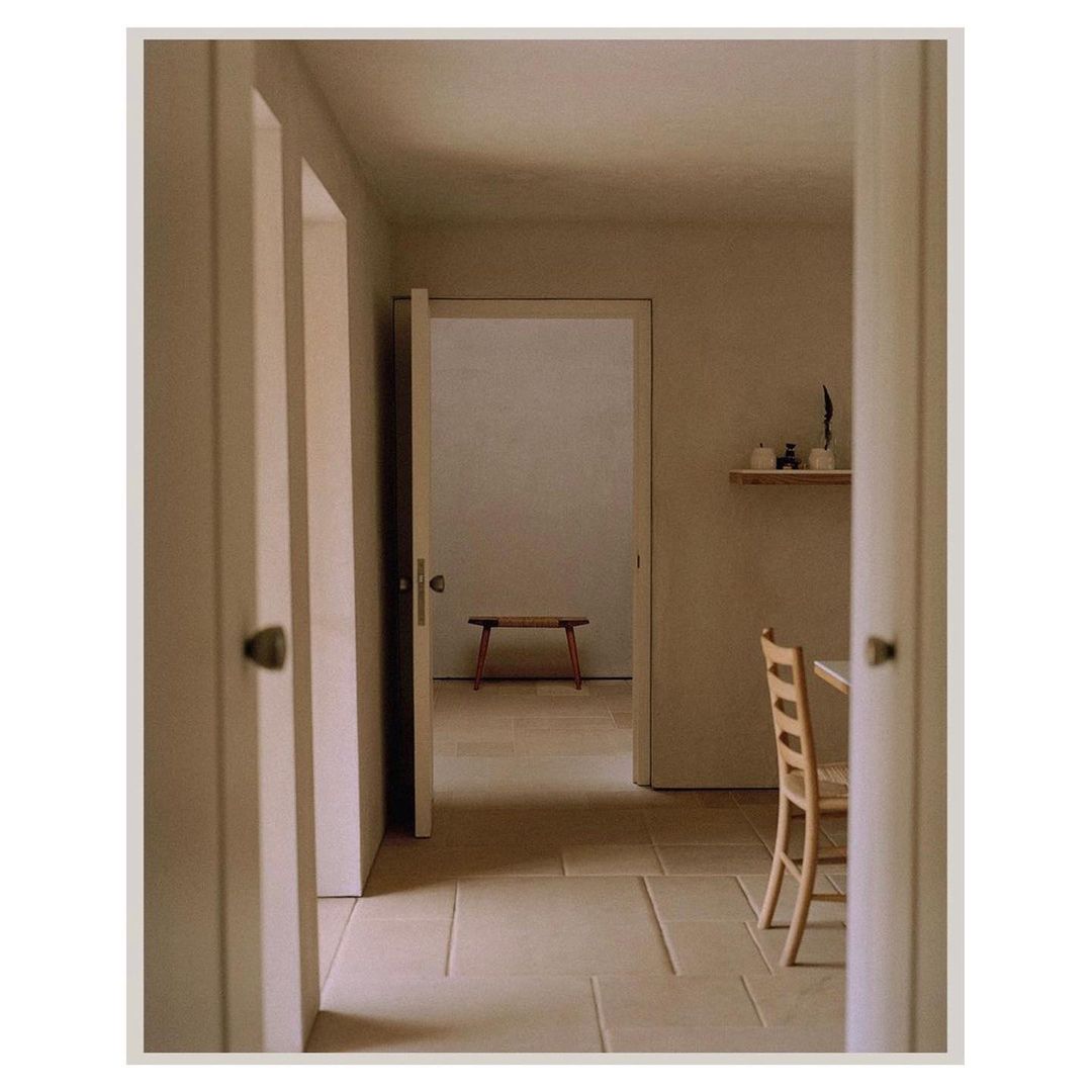 Minimalist interior with open doorways, a wooden bench, and a dining table with a chair, bathed in soft, neutral light on a tiled floor.