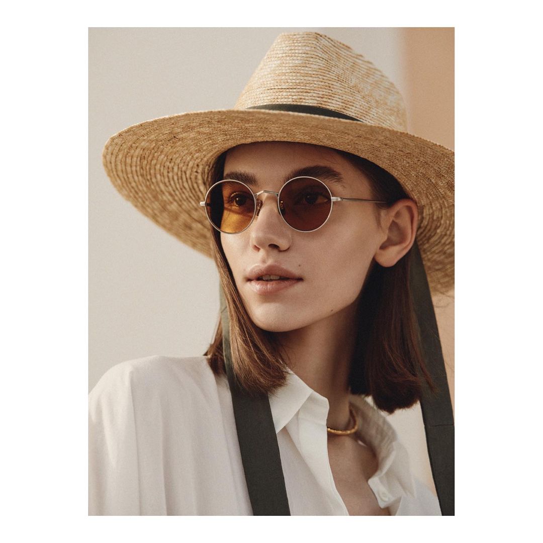 Woman wearing round sunglasses and a straw hat poses in a white button-down shirt with suspenders indoors in a fashion setting.