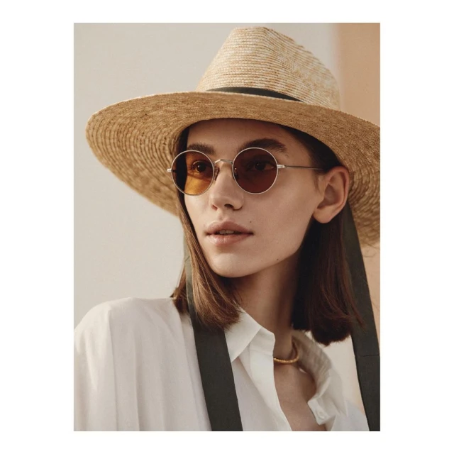 Woman wearing round sunglasses and a straw hat poses in a white button-down shirt with suspenders indoors in a fashion setting.