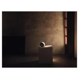 A sphere rests on a minimalist pedestal against a textured dark wall with light entering from a side window.