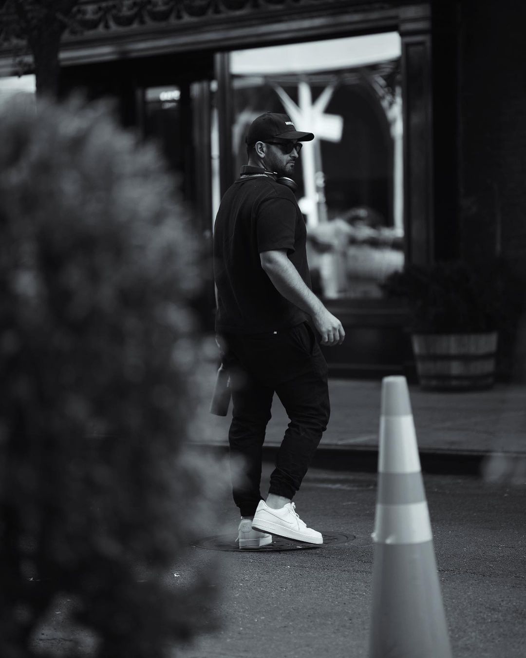 Black and white portrait of a man walking down a city street wearing a black shirt, pants, white sneakers and a black baseball cap.