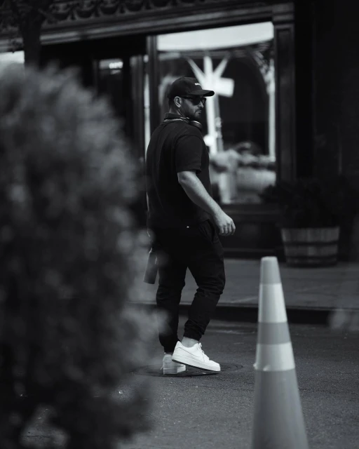 Black and white portrait of a man walking down a city street wearing a black shirt, pants, white sneakers and a black baseball cap.