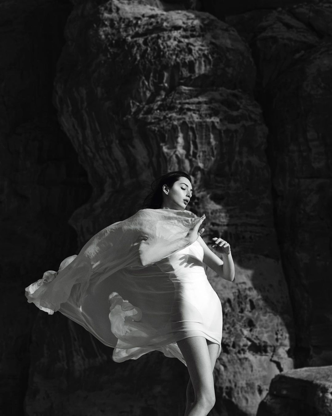 Black and white portrait of a woman in a light dress and flowing wrap posing against large rock formations.