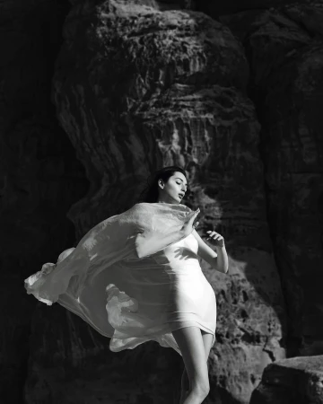 Black and white portrait of a woman in a light dress and flowing wrap posing against large rock formations.
