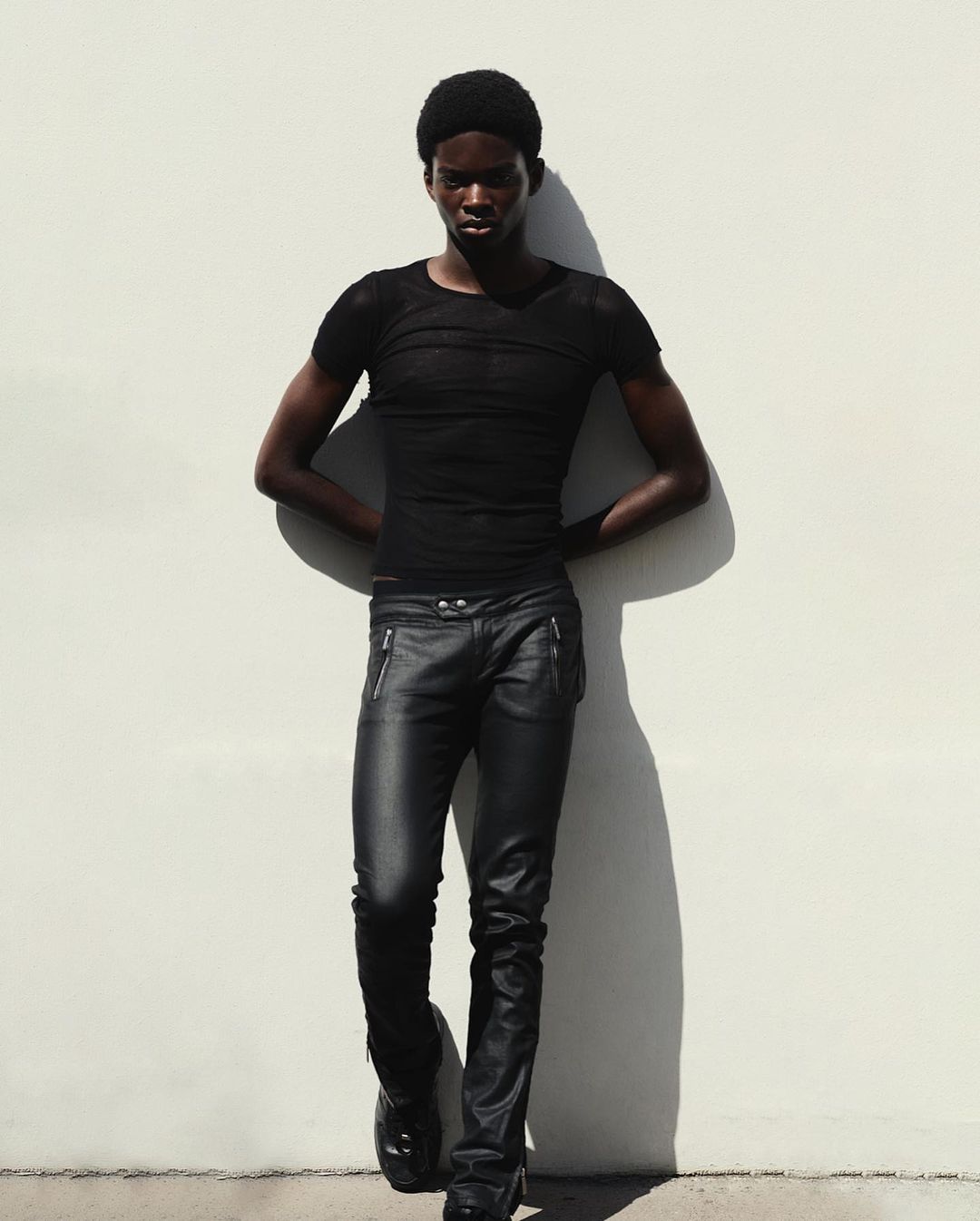 Young Black male fashion model posing in black top and leather pants against a white wall with dark shadows and high contrast.
