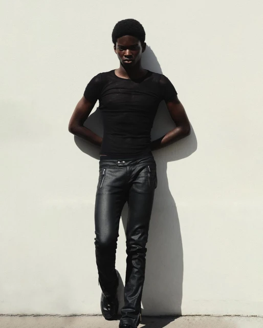 Young Black male fashion model posing in black top and leather pants against a white wall with dark shadows and high contrast.