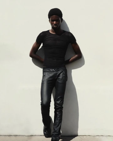 Young Black male fashion model posing in black top and leather pants against a white wall with dark shadows and high contrast.