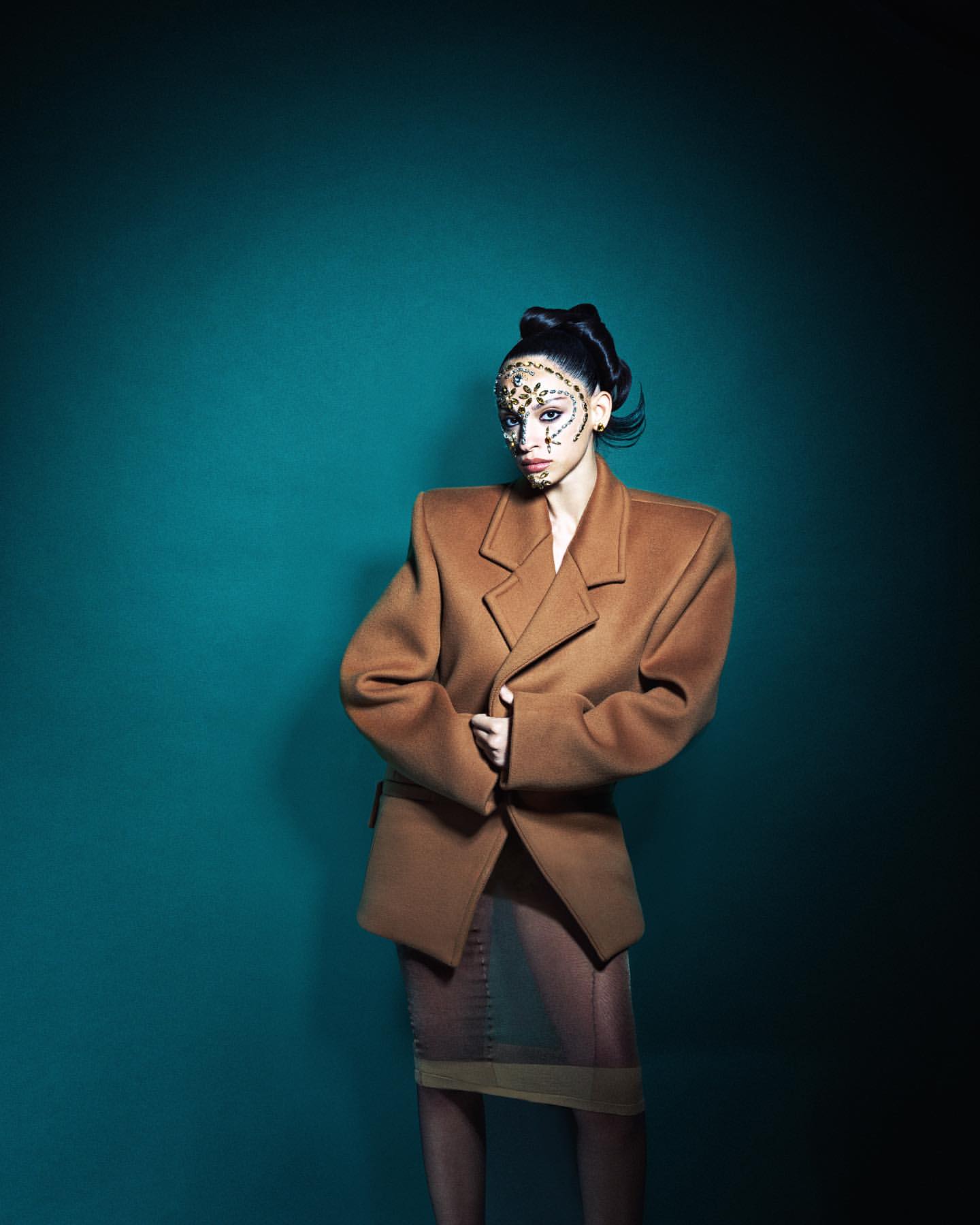 Fashionable woman in camel blazer with unique patterned makeup stands against a teal backdrop in a studio setting.