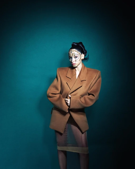 Fashionable woman in camel blazer with unique patterned makeup stands against a teal backdrop in a studio setting.