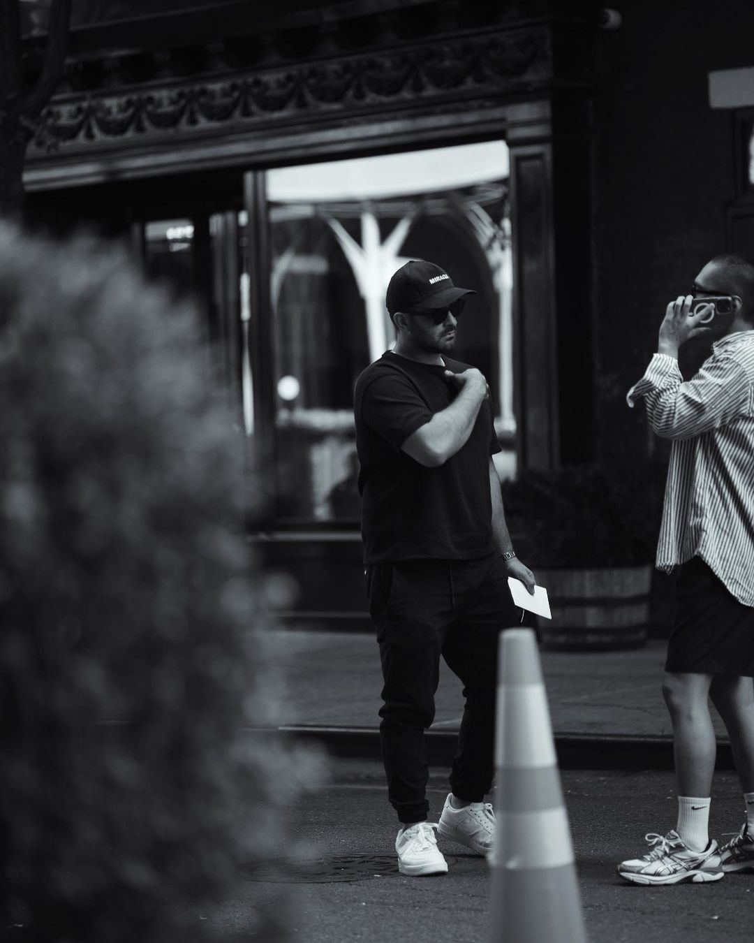Man wearing a dark t-shirt and baseball cap standing outdoors next to another person holding a phone, shot by Rayan Ayash.